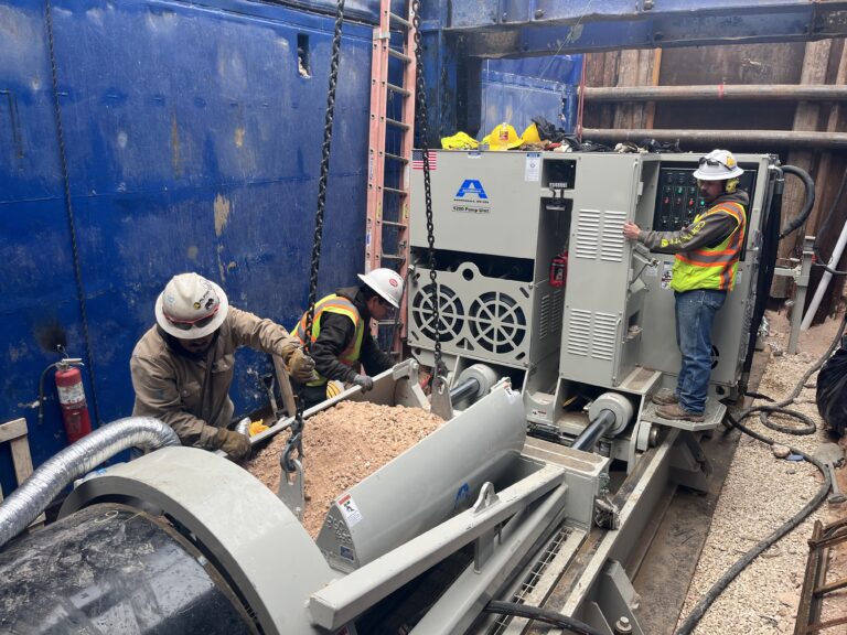 Workers operate pipe jacking equipment inside a trench box during a trenchless construction project.